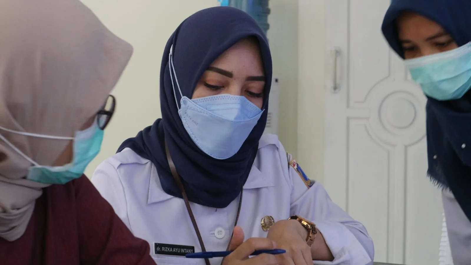 Seorang petugas RS Anggrek Mas berkerudung mencatat di meja kerja saat kunjungan validasi pengisian SIMATNEO, dengan dua rekannya berdiri di sekitarnya.
