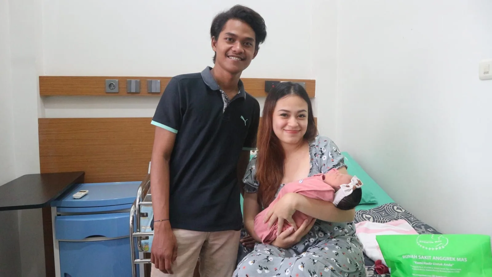 Sepasang suami istri berfoto di kamar perawatan; sang ibu duduk di tempat tidur sambil menggendong bayi yang baru lahir.