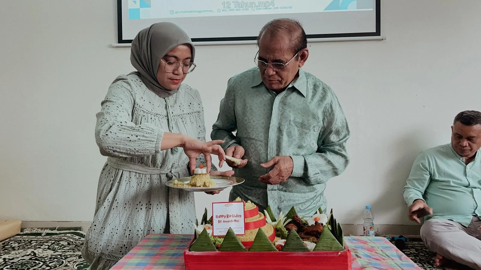 Seorang karyawati berkerudung dan seorang pria berkemeja hijau memotong tumpeng bersama pada tasyakuran ulang tahun ke-12 RS Anggrek Mas.