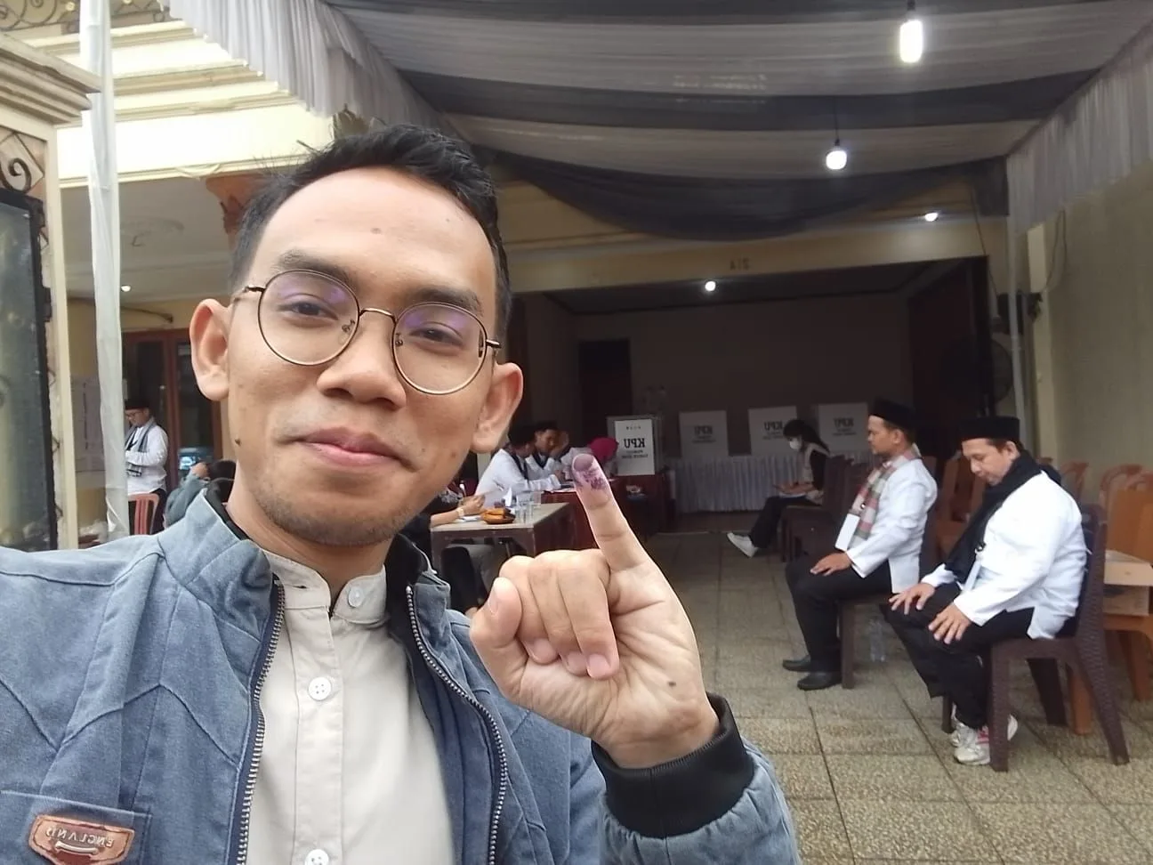 Seorang karyawan RS Anggrek Mas menunjukkan jari kelingking bertinta ungu seusai mencoblos di tempat pemungutan suara, dengan bilik suara KPU dan pemilih lain yang sedang menunggu di latar belakang.