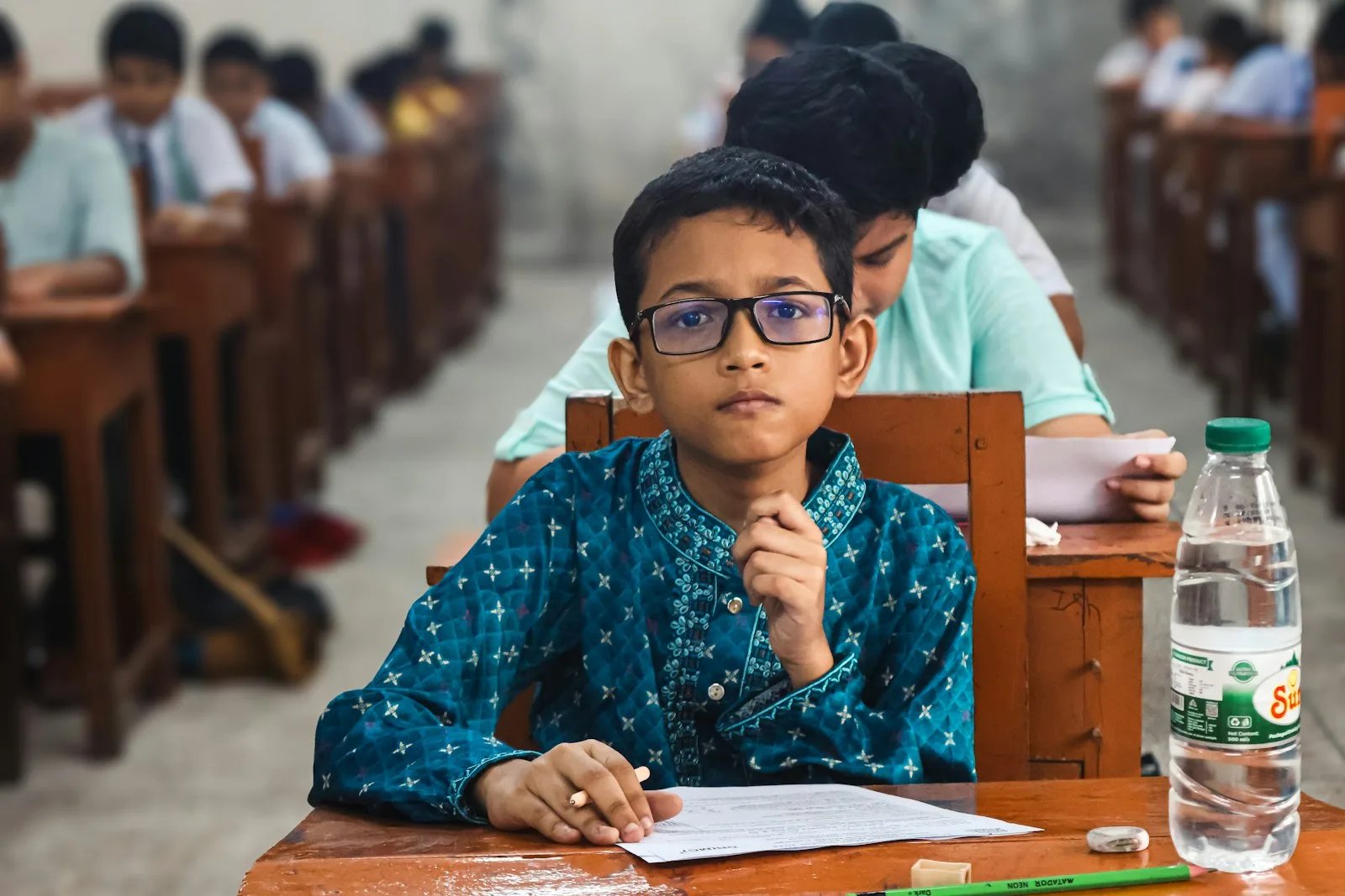 Seorang anak laki-laki berkacamata duduk di bangku kelas sambil mengerjakan tugas di atas kertas.