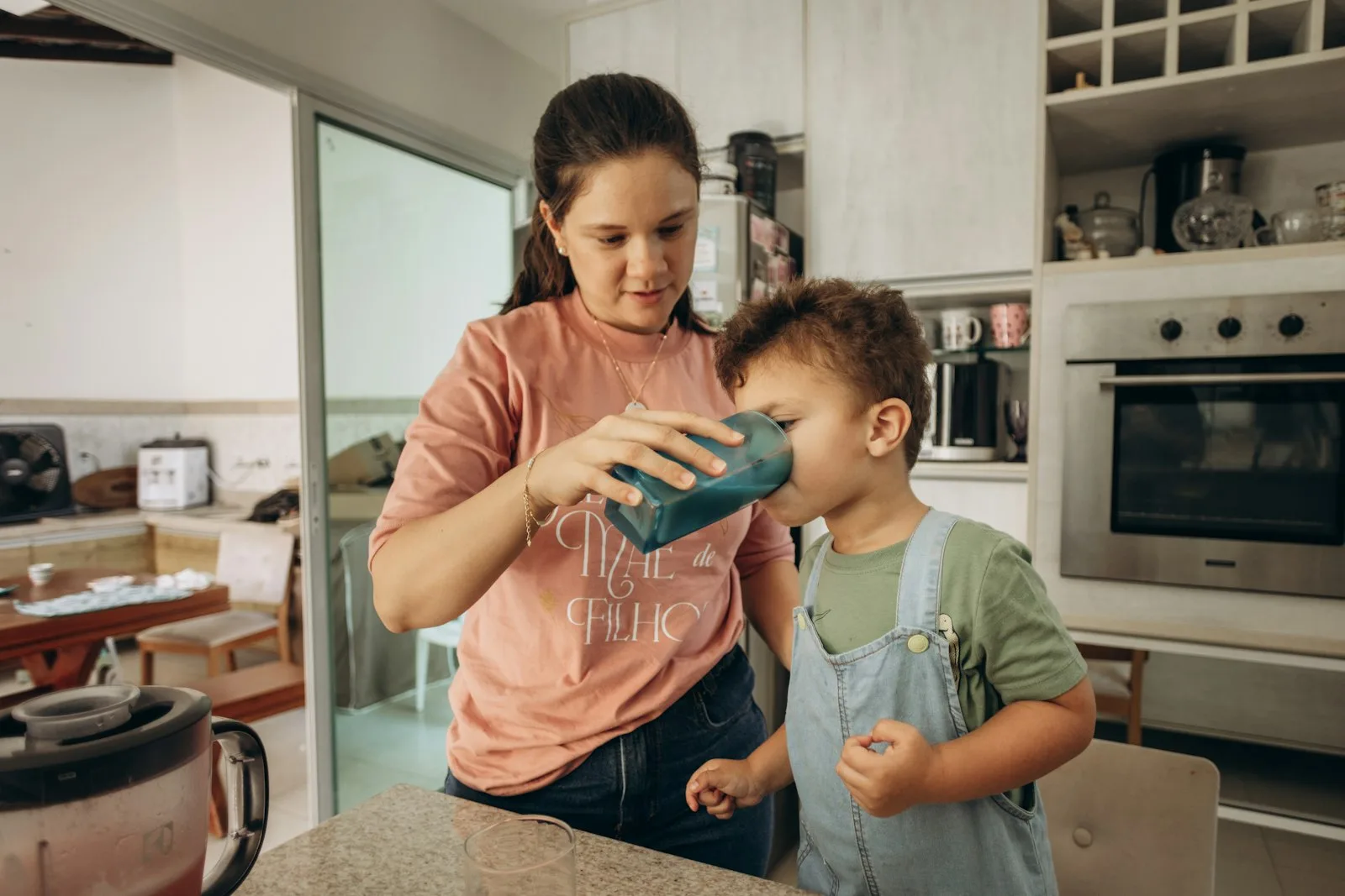 Seorang ibu memberikan minuman kepada anak balitanya di dapur rumah.